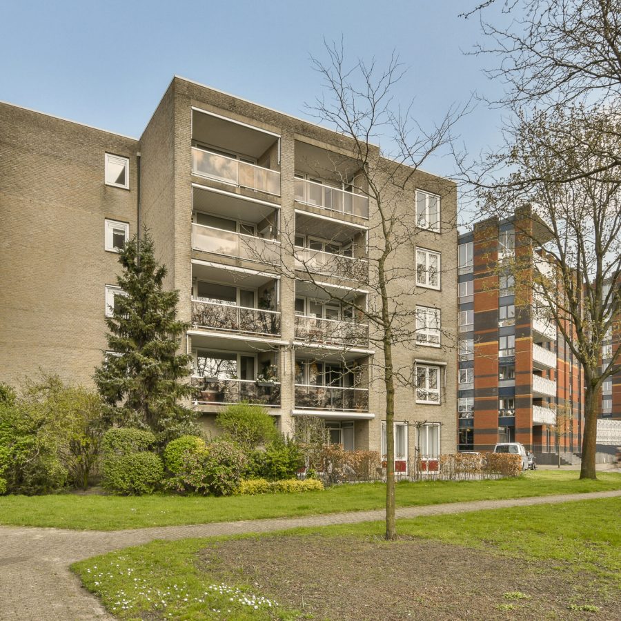 an apartment building with trees and bushes in the foreground area, on a clear blue sky day that is very sunny